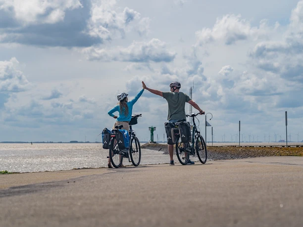 Emden-Außenhafen_01-CC_BY_SA-MaximilianSemsch_EmsRadweg.jpg Zwei Radfahrer am Hafen heben die Hände zum High-Five, im Hintergrund erstreckt sich das Meer.
