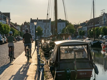 Weener-Alter Hafen_01-CC_BY_SA-MaximilianSemsch_EmsRadweg.jpg Radfahrer auf Kopfsteinpflasterstraße neben historischen Booten im Alten Hafen von Weener bei Sonnenuntergang.