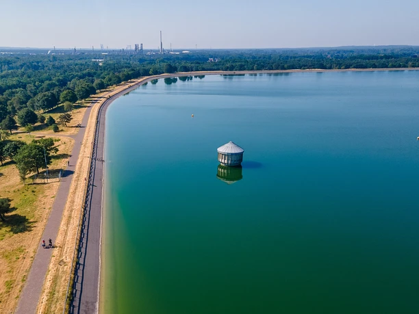 Geeste-Speichersee_01-CC_BY_SA-MaximilianSemsch_EmsRadweg.jpg Geeste-Speichersee mit Radweg, klarer Himmel, Wasser, vereinzeltes Gebäude im Wasser, im Hintergrund Industrie.