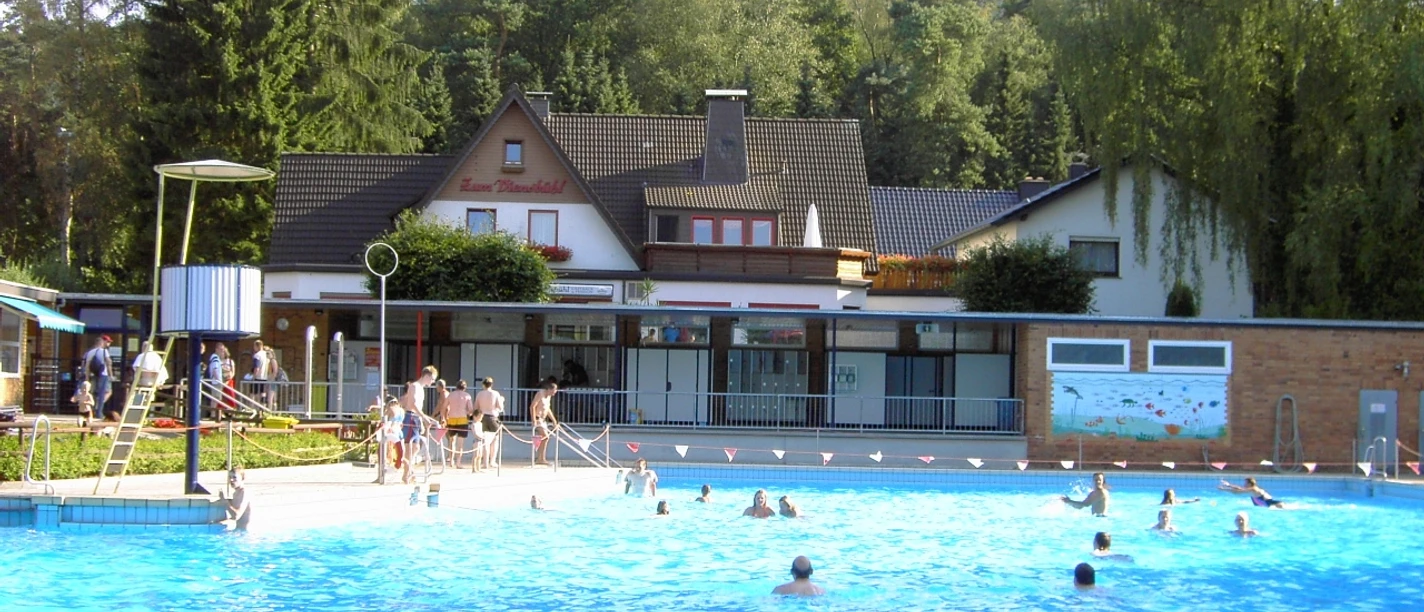 Das Freibad 2004.JPG Öffentliches Freibad mit Schwimmern, umgeben von Bäumen und Gebäuden unter einem klaren Himmel.