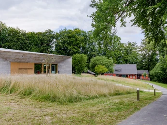 Teutoburger Wald / Museum Peter August Böckstiegel / Werther (Westf.) / Foto: M. Wallenfang Moderner Museumsgebäude mit großen Fenstern, umgeben von Rasen und Bäumen; traditionelle Holzhäuser im Hintergrund.