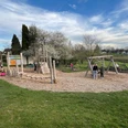 Spielplatz Ein Spielplatz in einer grünen Parklandschaft mit Kindern, die auf Holzspielgeräten spielen und Eltern, die zuschauen.