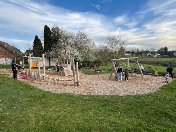 Spielplatz Ein Spielplatz in einer grünen Parklandschaft mit Kindern, die auf Holzspielgeräten spielen und Eltern, die zuschauen.