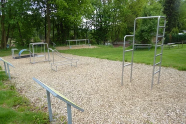 Fitnessparcours Ein Outdoor-Fitnesspark mit mehreren Geräten auf Holzschnitzelboden, umgeben von grünen Bäumen.