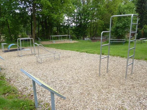 Fitnessparcours Ein Outdoor-Fitnesspark mit mehreren Geräten auf Holzschnitzelboden, umgeben von grünen Bäumen.