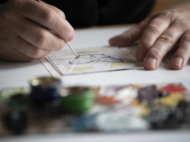 Hinterglasmalerei Dichtl Eine Hand malt mit Pinsel auf Glas, bunte Farben im Vordergrund.