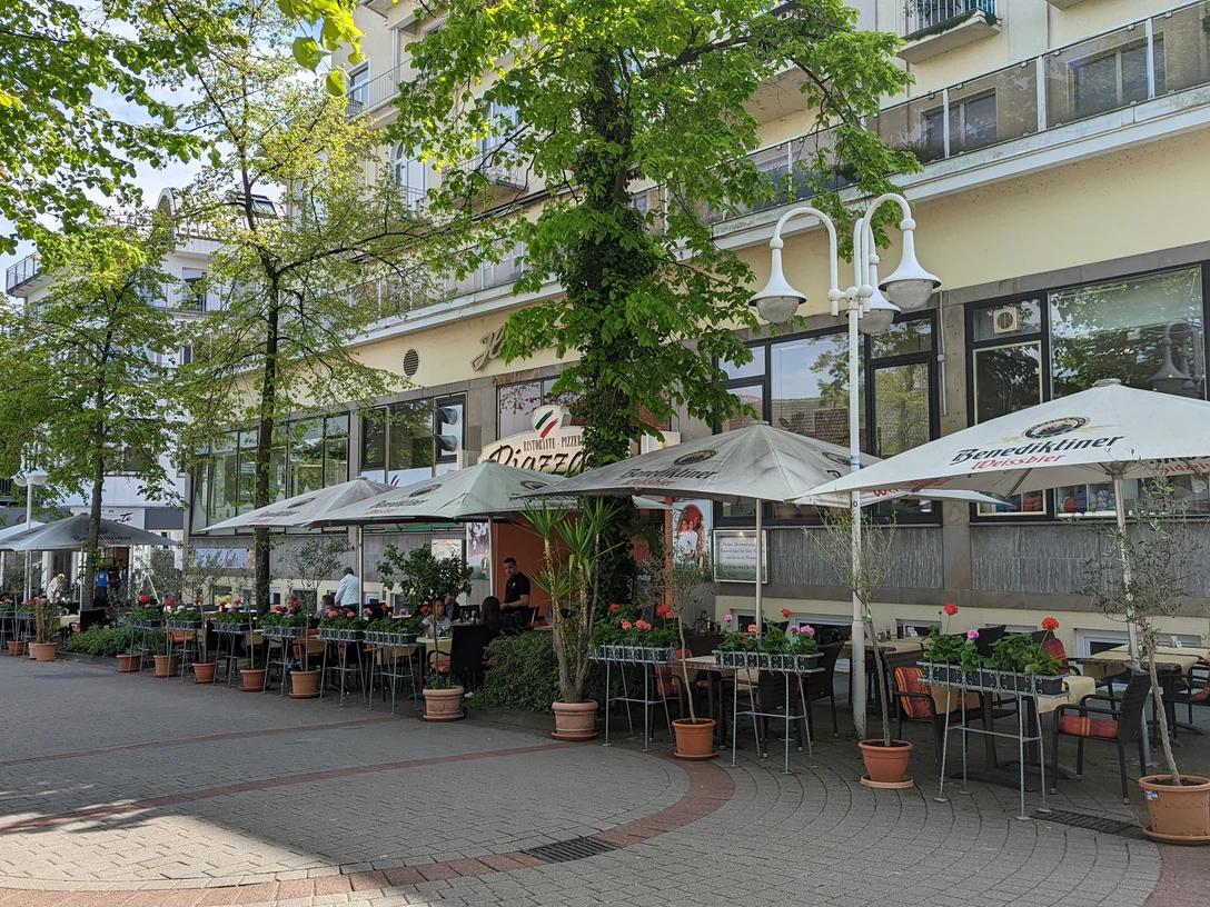 Ristorante Pizzeria Piazza.jpg Auf dem Bild ist die einladende Außenansicht des Restaurants zu sehen, umgeben von üppigen, grünen Bäumen. Gemütliche Sitzgelegenheiten laden dazu ein, die frische Luft zu genießen und im Freien zu speisen.