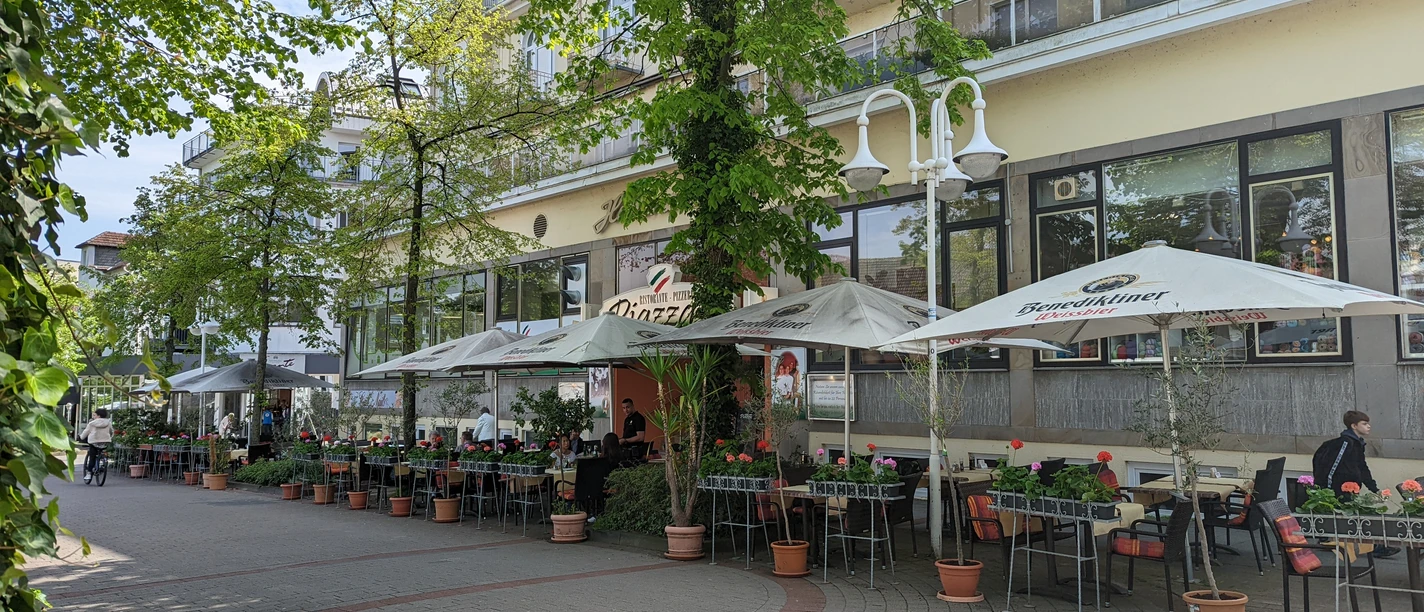 Ristorante Pizzeria Piazza.jpg Auf dem Bild ist die einladende Außenansicht des Restaurants zu sehen, umgeben von üppigen, grünen Bäumen. Gemütliche Sitzgelegenheiten laden dazu ein, die frische Luft zu genießen und im Freien zu speisen.