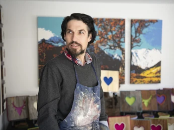 Alpenterieur Künstler in Atelier mit bunten Bildern im Hintergrund, trägt Arbeitsbekleidung.