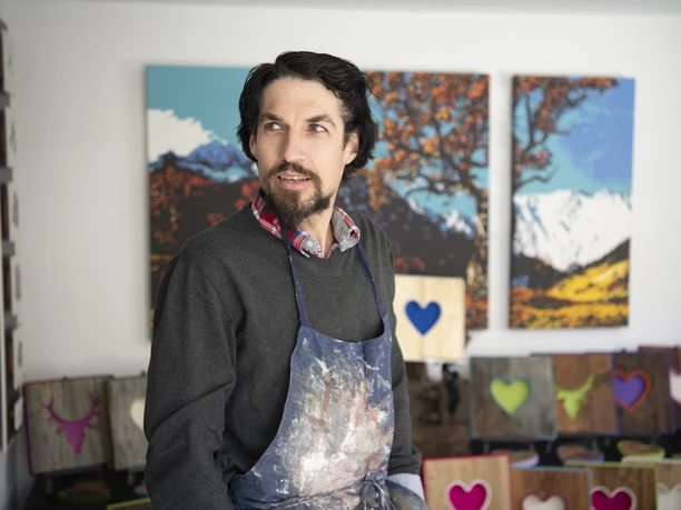 Alpenterieur Künstler in Atelier mit bunten Bildern im Hintergrund, trägt Arbeitsbekleidung.