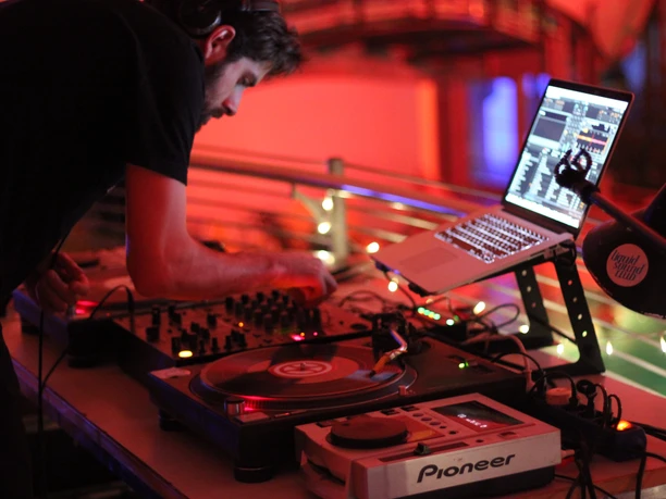 Liquid Sound Club Een DJ treedt op in een schemerige kamer met moderne apparatuur, omgeven door rood licht.