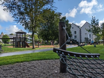 Spielplatz im Kurpark Grünanlage mit Spielplatz, Kletternetz im Vordergrund, Mehrfamilienhäuser und Erwachsene im Hintergrund.