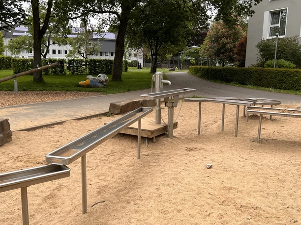 Wasserspielplatz im Kurpark Barrierefreier Spielplatz mit Sand und Edelstahl-Wasserspiel in einer grünen Parkanlage vor Gebäuden.