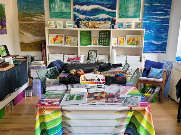 Atelier von Andrea Molière Ein bunter Künstlerraum mit Gemälden von Wellen und Blumen an den Wänden, ein Tisch mit Malutensilien und farbenfroher Tischdecke.