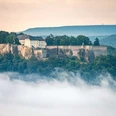 Festung Königstein Die historische Festung Königstein thront auf einem bewaldeten Hügel, umgeben von Nebel, unter einem sanft bewölkten Himmel.