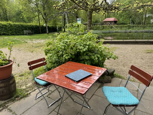 Café Alte Dombach - Außenansicht Ein rot lackierter Holztisch mit zwei Stühlen steht im grünen Garten vor üppigem Pflanzenwuchs.