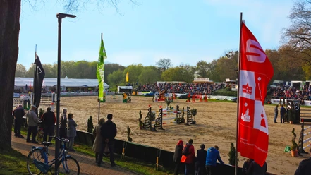 Sielturnier Das Bild zeigt eine große, sandige Turnierfläche mit mehreren Hindernissen und Reitern in Aktion. Im Vordergrund stehen Zuschauer am Rand, während im Hintergrund weiterer Zuschauertribünen, Bäume und Zelte zu sehen sind.