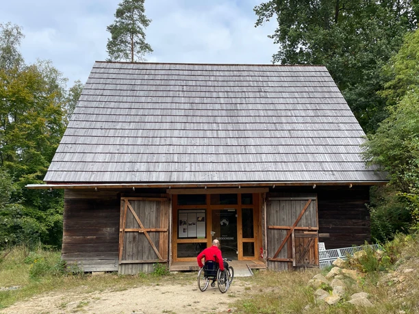 Een persoon in een rolstoel draagt een rode jas en nadert een houten gebouw met grote dubbele deuren, omringd door bomen.
