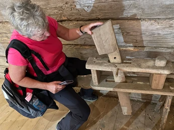 Blockscheune Waldhusche Eine Frau in einem pinken T-Shirt betrachtet ein altes hölzernes Werkzeug in einer rustikalen Holzhütte.