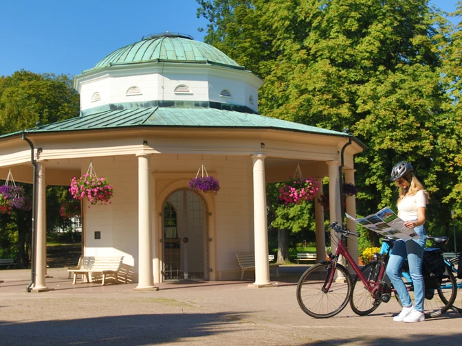 Ein Radfahrer mit Helm studiert eine Karte vor einem historischen Tempel mit Säulen und Blumenampeln.