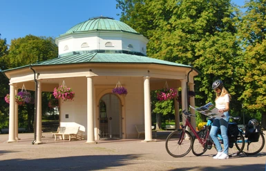 Radfahren_Brunnentempel_®GesUndTourismus Horn-Bad Meinberg GmbH.JPG Ein Radfahrer mit Helm studiert eine Karte vor einem historischen Tempel mit Säulen und Blumenampeln.