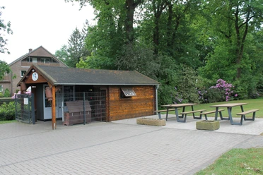 Laxtener Milchbüdchen in Lingen (Ems) ©Emsland Tourismus GmbH (10).JPG Kleines Holzhaus mit Milchautomaten und Sitzbänken im Grünen, umgeben von Bäumen und Blumen.