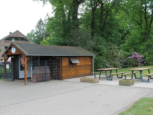 Laxtener Milchbüdchen in Lingen (Ems) ©Emsland Tourismus GmbH (10).JPG Kleines Holzhaus mit Milchautomaten und Sitzbänken im Grünen, umgeben von Bäumen und Blumen.