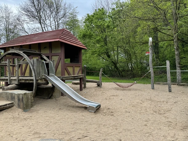 Café Alte Dombach - Spielplatz Spielplatz mit Holzhaus, Rutsche, Hängematte auf Sandboden, umgeben von Bäumen.