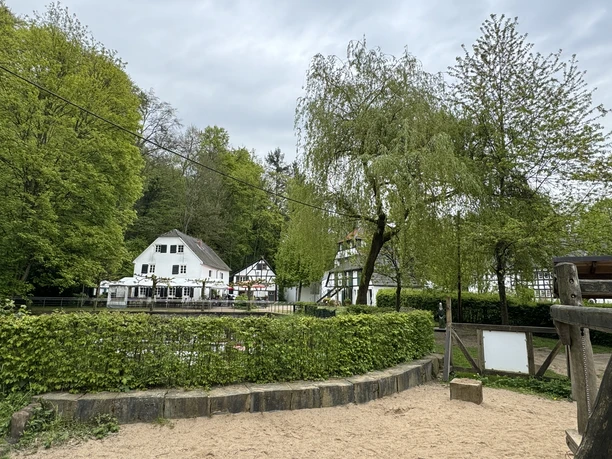 Café Alte Dombach - Ansicht Haus Ein malerisches Fachwerkhaus am Waldrand, umgeben von üppigem Grün und einem Sandspielplatz.