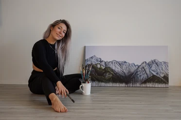 Ayla Ginsberg Künstlerin sitzt mit Pinsel in der Hand vor einem Berggemälde, lächelnd auf dem Boden.