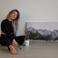 Ayla Ginsberg Künstlerin sitzt mit Pinsel in der Hand vor einem Berggemälde, lächelnd auf dem Boden.