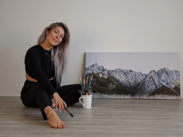 Ayla Ginsberg Künstlerin sitzt mit Pinsel in der Hand vor einem Berggemälde, lächelnd auf dem Boden.