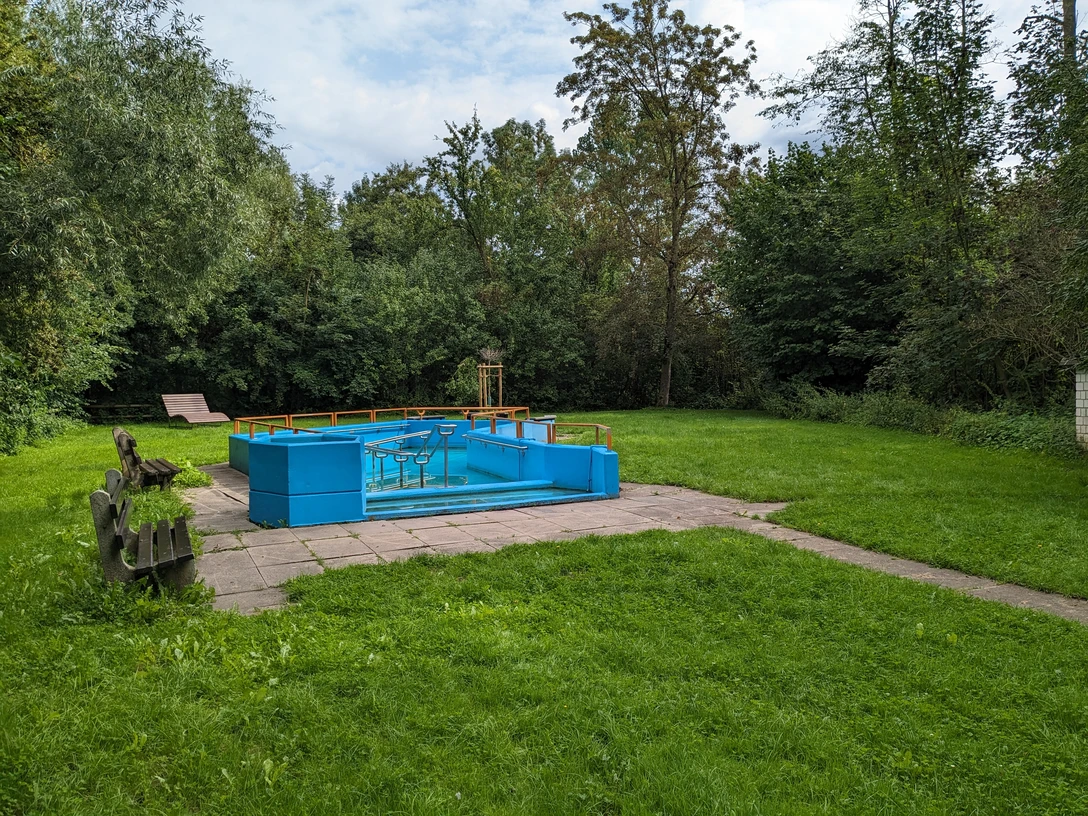 Blaues Kneipptretbecken, umgeben von Pflastersteinen und Rasen, linksseitig zwei Holzbänke, im Hintergrund Bäume und eine Ruhebank