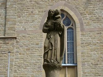 Statue an der Kirche Maria-Statue vor einer Kirche aus Sandstein, sie hält ein Kind auf dem Arm. Im Hintergrund Kirchenwand.
