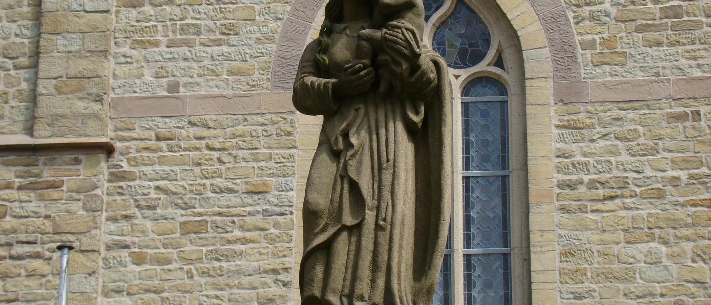 Statue an der Kirche Maria-Statue vor einer Kirche aus Sandstein, sie hält ein Kind auf dem Arm. Im Hintergrund Kirchenwand.