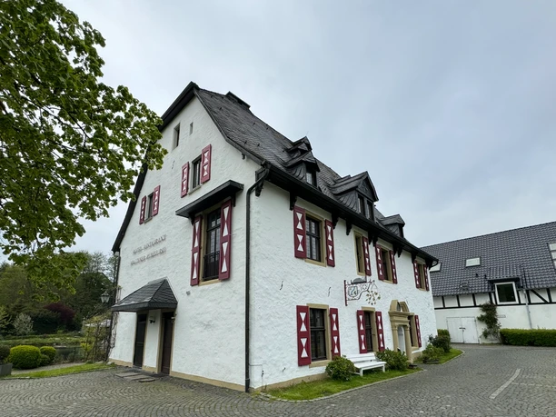 Außenansicht Fachwerkhaus mit weißen Wänden, roten Fensterläden und Schieferdach, umgeben von gepflastertem Hof.