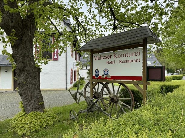 Schild Malteser Komturei: Historisches Gebäude mit beschattetem Schild, umgeben von grüner Natur.