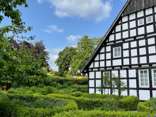 Heimathaus Wehdem Fachwerkhaus mit gepflegtem Garten und Hecken, unter blauem Himmel mit Wolken im Hintergrund.