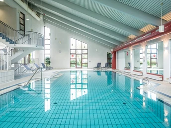 Trans World Hotel Kranichhöhe Innenansicht eines modernen, lichtdurchfluteten Hallenbads mit klarem Wasser und Liegestühlen.