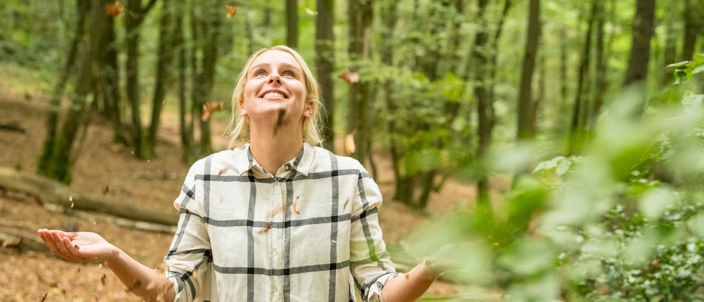Waldbaden Frau in kariertem Hemd genießt lachend einen Herbstspaziergang im Wald unter fallendem Laub.