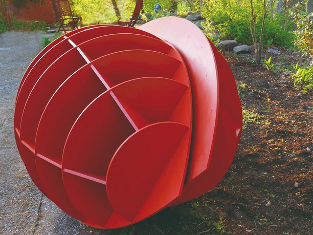 Harald a. Finke Modell Pflanzendenkmal-Chloroplast-Mitochondrium 2022 Das Bild zeigt eine kunstvolle, rot bemalte Skulptur im Freien, die kugelförmig gestaltet ist und aus mehreren Segmenten besteht. Sie liegt auf einem gepflasterten Weg und wird von grüner Vegetation umrahmt.