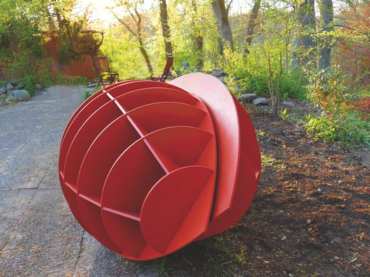 Harald a. Finke Modell Pflanzendenkmal-Chloroplast-Mitochondrium 2022.jpg Das Bild zeigt eine kunstvolle, rot bemalte Skulptur im Freien, die kugelförmig gestaltet ist und aus mehreren Segmenten besteht. Sie liegt auf einem gepflasterten Weg und wird von grüner Vegetation umrahmt.