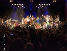 Große Menschenmenge feiert ausgelassen auf einer Bühne unter bunten Lichtern in einem Veranstaltungsraum.