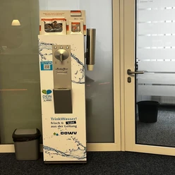 Trinkwasserspender im Rathaus_Manuela Baumhöfer.jpg Silberner Trinkwasserspender mit Bechern und Infomaterial im Eingangsbereich eines Büros.
