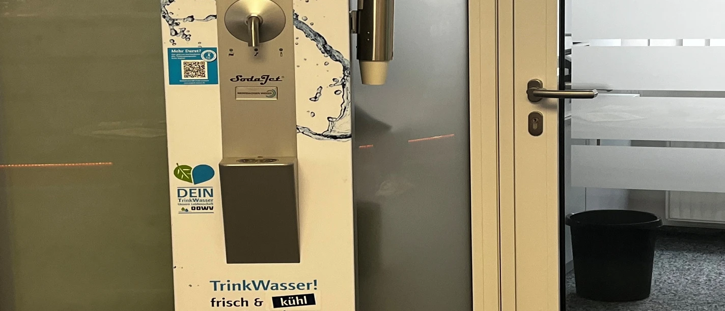 Trinkwasserspender im Rathaus_Manuela Baumhöfer.jpg Silberner Trinkwasserspender mit Bechern und Infomaterial im Eingangsbereich eines Büros.