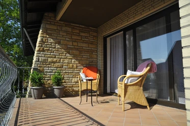 Hotel zur Herzklinik - Balkon.JPG Sonniger Balkon mit Rattansesseln und Tisch, begrenzt von einem geschwungenen Eisenzaun.