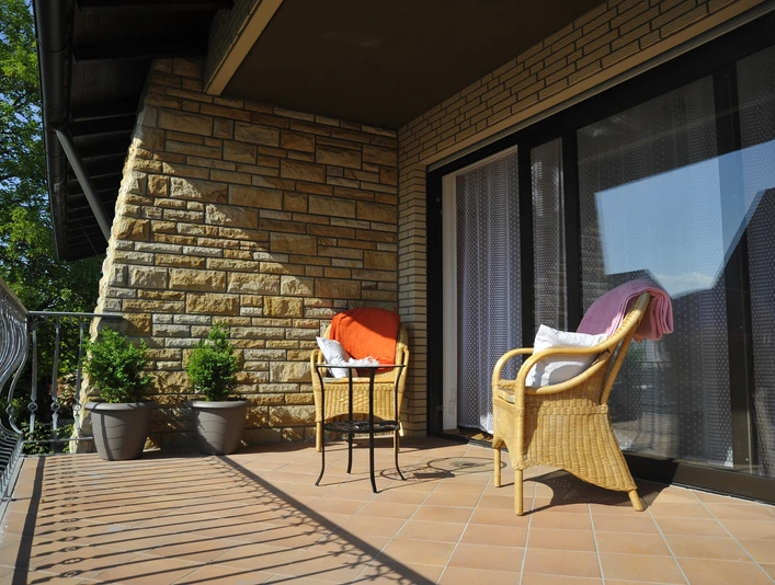 Hotel zur Herzklinik - Balkon.JPG Sonniger Balkon mit Rattansesseln und Tisch, begrenzt von einem geschwungenen Eisenzaun.