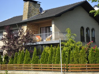Hotel zur Herzklinik - Außenansicht.JPG Zweistöckiges Haus mit Backsteinfassade, Balkon mit Stühlen, Ziegelkamin und üppiger grüner Bepflanzung.