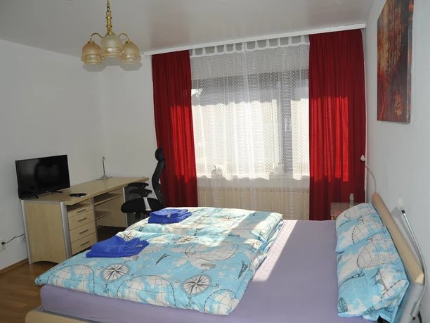 Hotel zur Herzklinik - Zimmer 1.JPG Helles Zimmer mit Doppelbett, Schreibtisch, TV, Leuchte und Fenster mit rotem Vorhang.