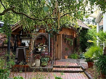 Gartenhäuschen Angelika und Holger Ihler.jpg Ein charmantes Gartenhäuschen aus Backstein mit mediterranen Pflanzen umgeben von Bäumen.